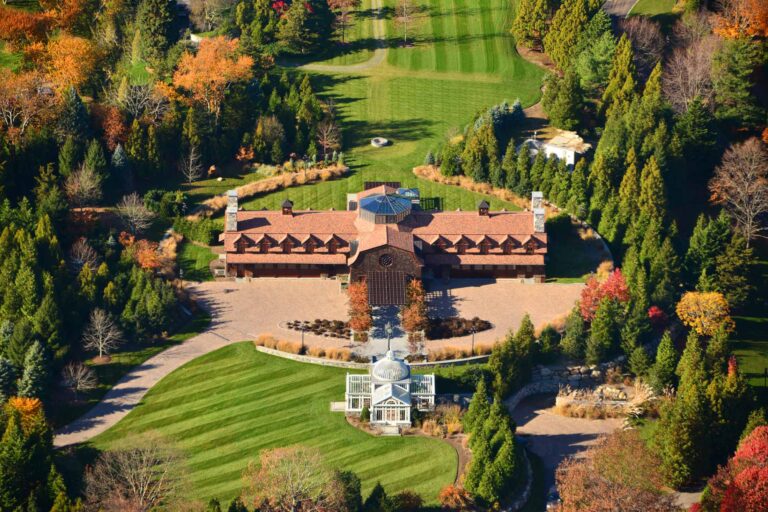 Aerial view of a Head of the Harbor Shingle-style entertainment retreat with a circular arrival court, glass conservatory, formal lawns, gardens, and mature trees.