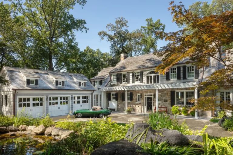 Front exterior of historic Sands Point Family Residence with stone facade, slate roof, pergola entry, and landscaped garden pond