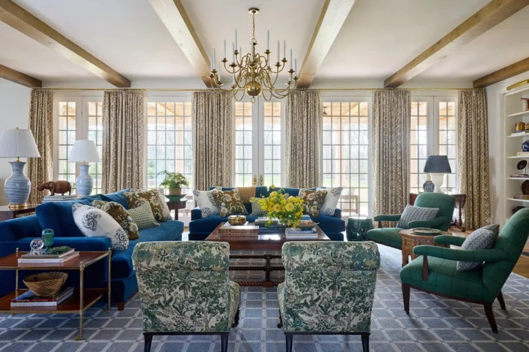 Wide living room view with multiple windows, exposed ceiling beams, blue sofa, green chairs, and patterned carpet