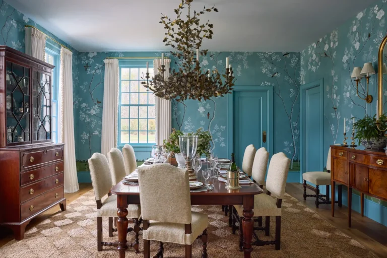 Dining room with hand-painted blue wallpaper panels, antique wood table, upholstered chairs, chandelier, and tall window