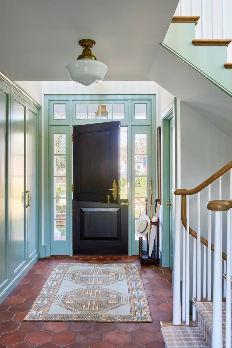 Side entry hall with green paneled door, patterned terracotta tile floor, stair rail, and light painted millwork