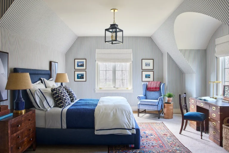 Bedroom with blue bed, patterned wallpaper, vaulted ceiling, dormer windows, and upholstered sitting chair