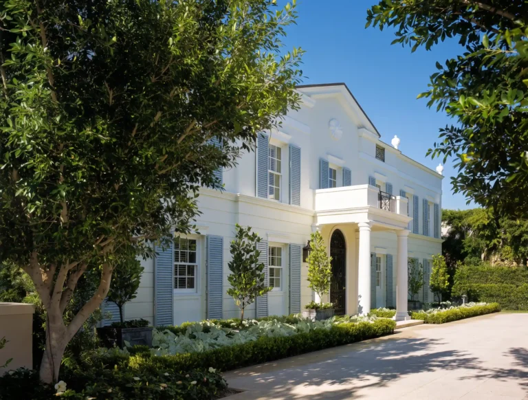 Front exterior of a white Regency-style Palm Beach residence with an entry portico, green shutters, manicured plantings, and a shaded drive.