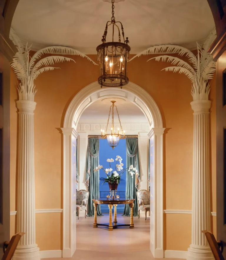 Palm Beach foyer with arched openings, palm-inspired columns, lantern pendant, warm wall color, and an axial view into an evening-lit room beyond.