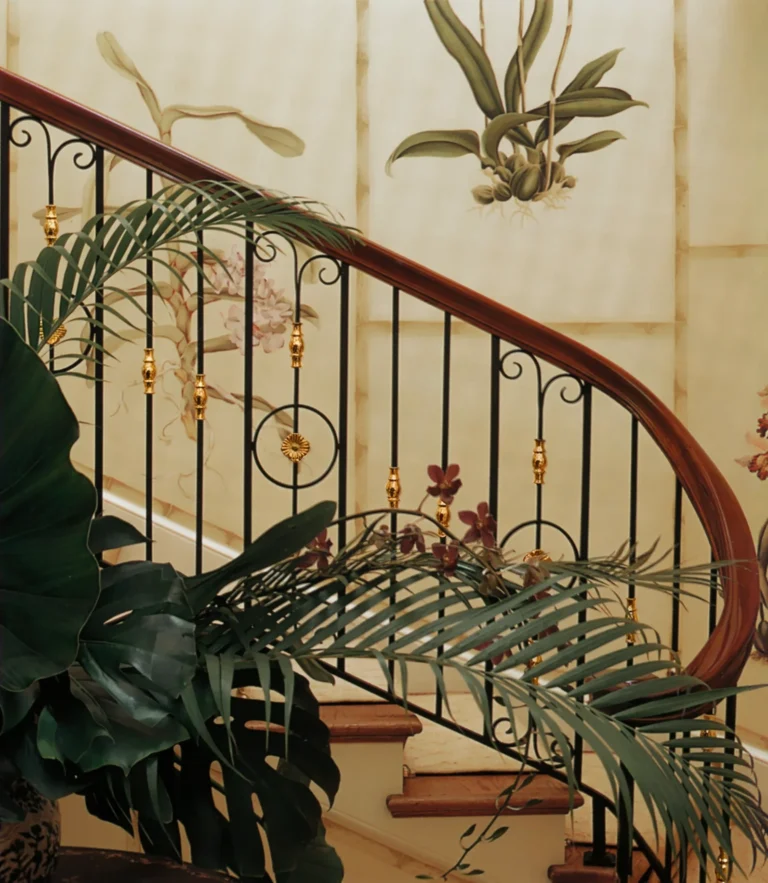 Curved stair in a Palm Beach residence with decorative iron railing, wood handrail, botanical wall detail, and tropical plantings.