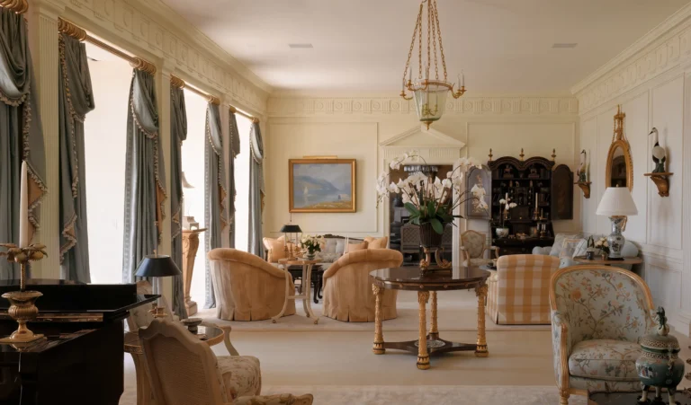 Second view of a formal living room with tall windows, patterned draperies, chandelier, seating groups, side tables, and classical wall detailing.