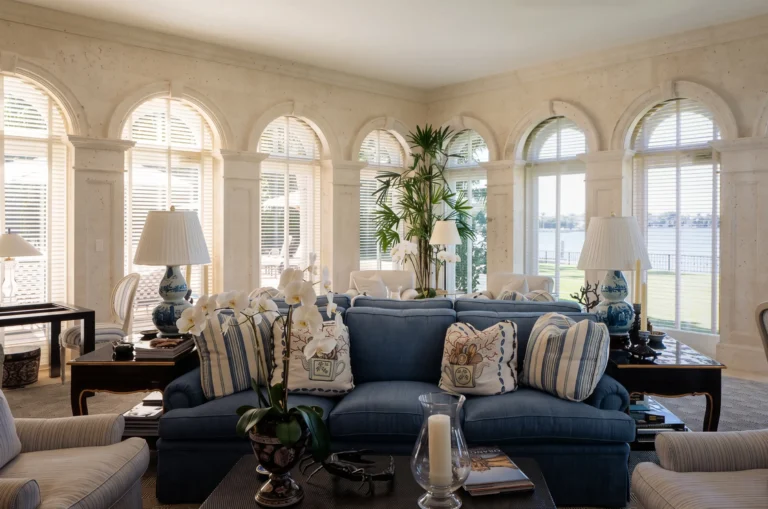 Light-filled family room with arched windows, blue sofa, pale lounge chairs, palm planting, water views, and classical wall detailing.