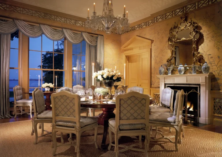 Formal dining room with chandelier, round dining table, upholstered chairs, fireplace, gilt mirror, draperies, and evening garden views.