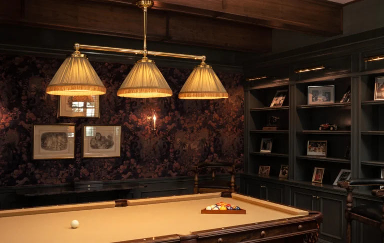 Billiards room with dark academia built-in shelving, patterned wallcovering, framed art, pendant lights over a pool table, and warm wood trim.
