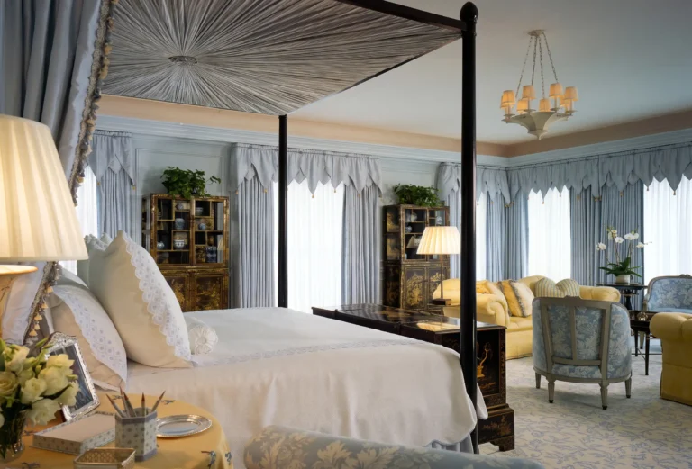 Palm Beach bedroom with four-poster bed, pale bedding, patterned ceiling treatment, upholstered seating, draperies, and garden light.