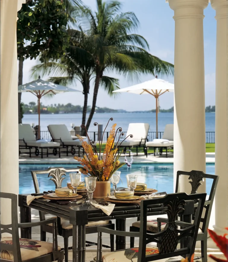 Columned porch with outdoor dining table, classical columns, poolside seating, palm trees, umbrellas, and water views in Palm Beach.