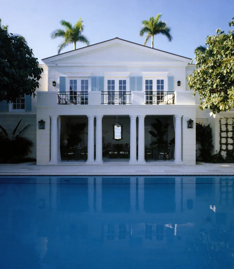 Swimming pool facing the white rear elevation of a Regency-style Palm Beach residence with French doors, columns, and balcony above.