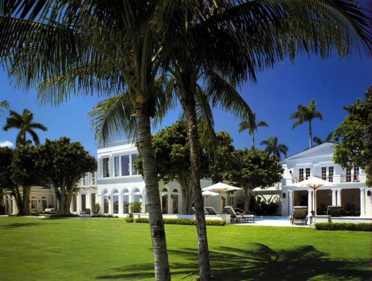 Rear lawn of a white Palm Beach Regency residence with palm trees, arched loggia, cabana-like wing, and manicured garden landscape.