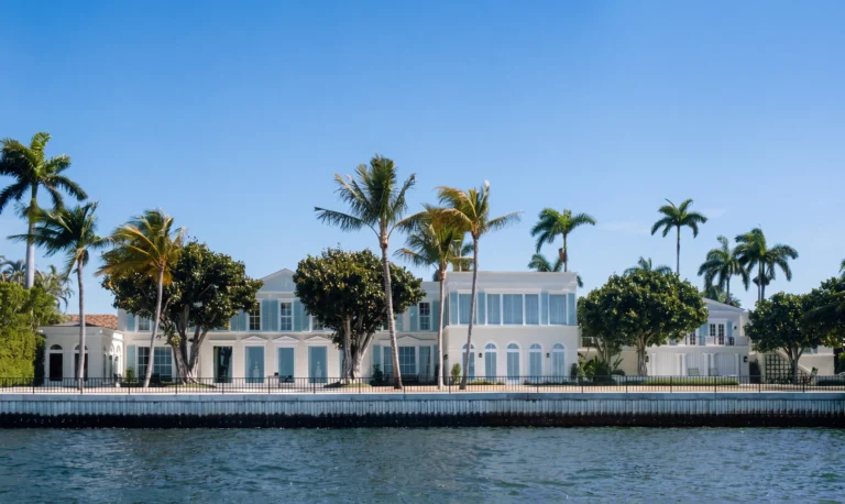 Waterfront view of a white Palm Beach Regency residence with palm trees, sea wall, rear façade, and formal lawn along the water.