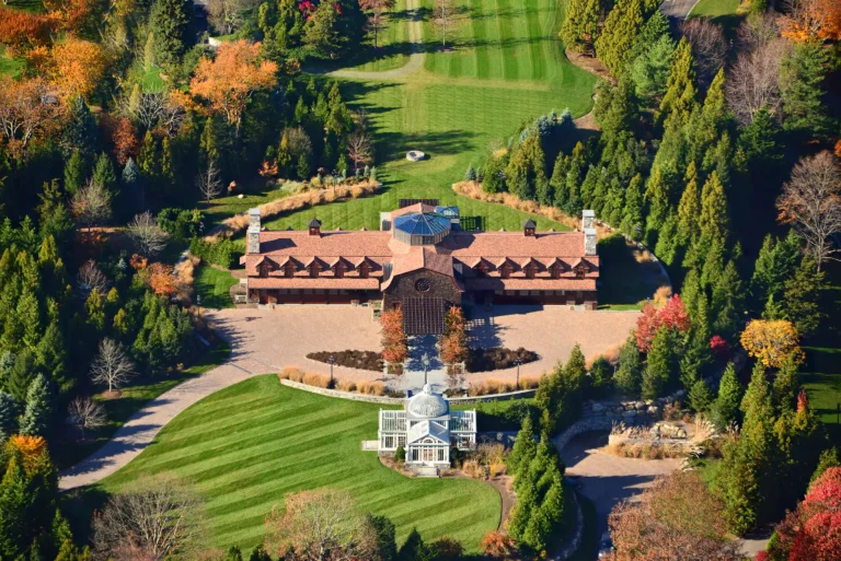 Aerial view of a Head of the Harbor Shingle-style entertainment retreat with a circular arrival court, glass conservatory, formal lawns, gardens, and mature trees.