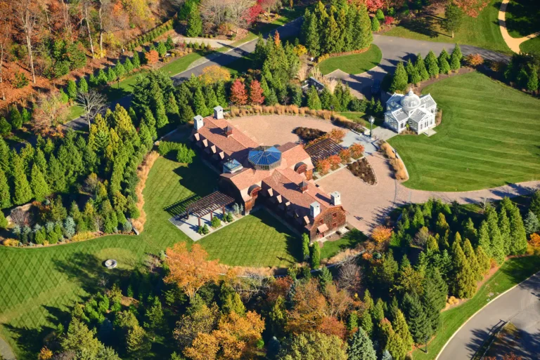 High aerial view of a Shingle-style estate retreat in Head of the Harbor with red roofs, formal drive, conservatory, broad lawns, rose garden, and autumn trees.