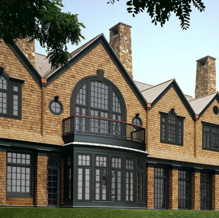 Rear lawn view of a waterfront Shingle Style residence with steep gables, large arched windows, brick chimneys, and French doors opening to the landscape.